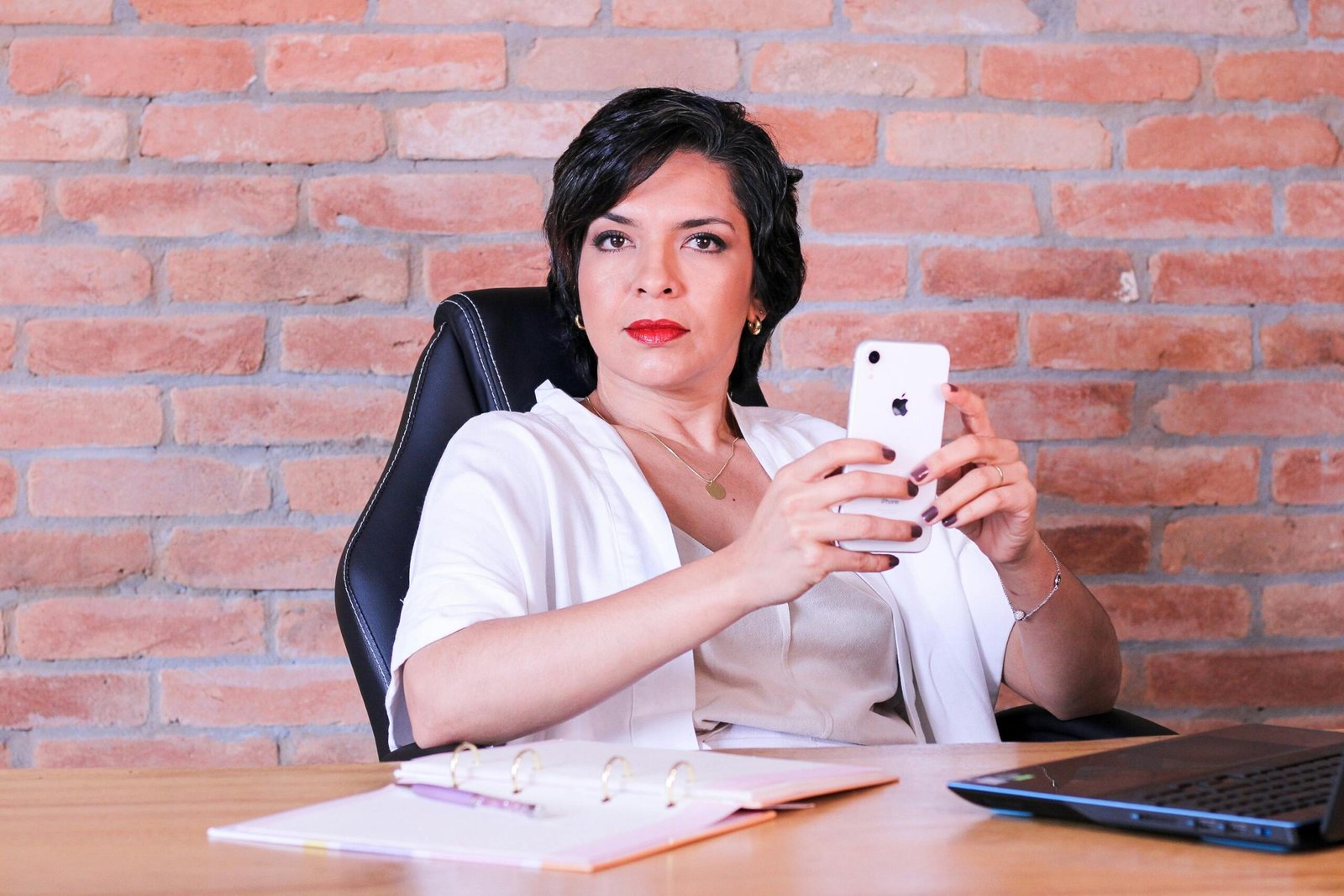 Professional woman using smartphone at desk with brick wall backdrop, representing remote work.